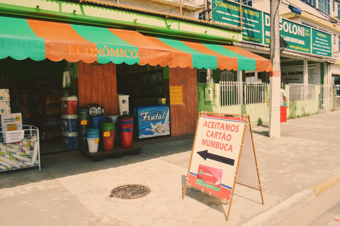 Foto de uma placa em frente a uma loja, onde se lê 'Aceitamos Cartão Mumbuca'.